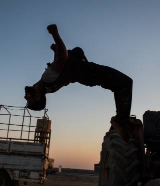 A fighter of the YPG doing a backflip from a vehicle.